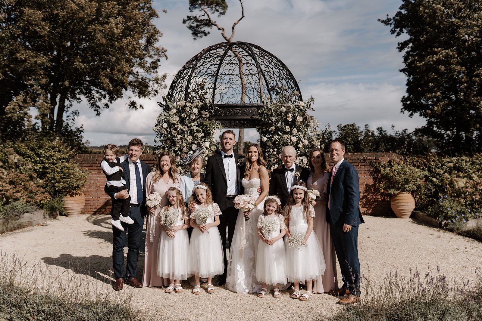 Wedding party posing outdoors with floral arbor in background, featuring bride, groom, family members, and flower girls.
