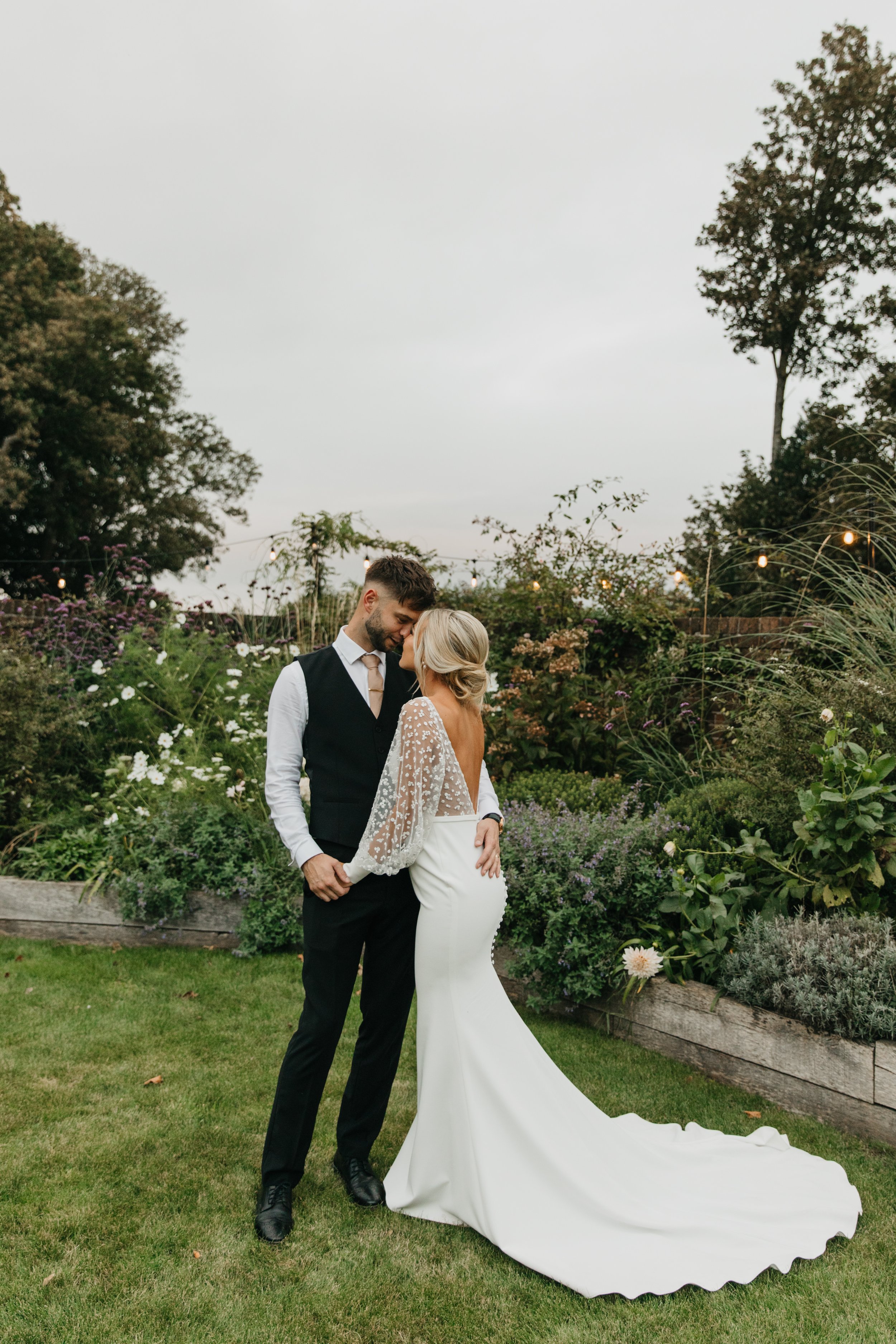 Bride and groom embracing in garden setting, wearing wedding attire.