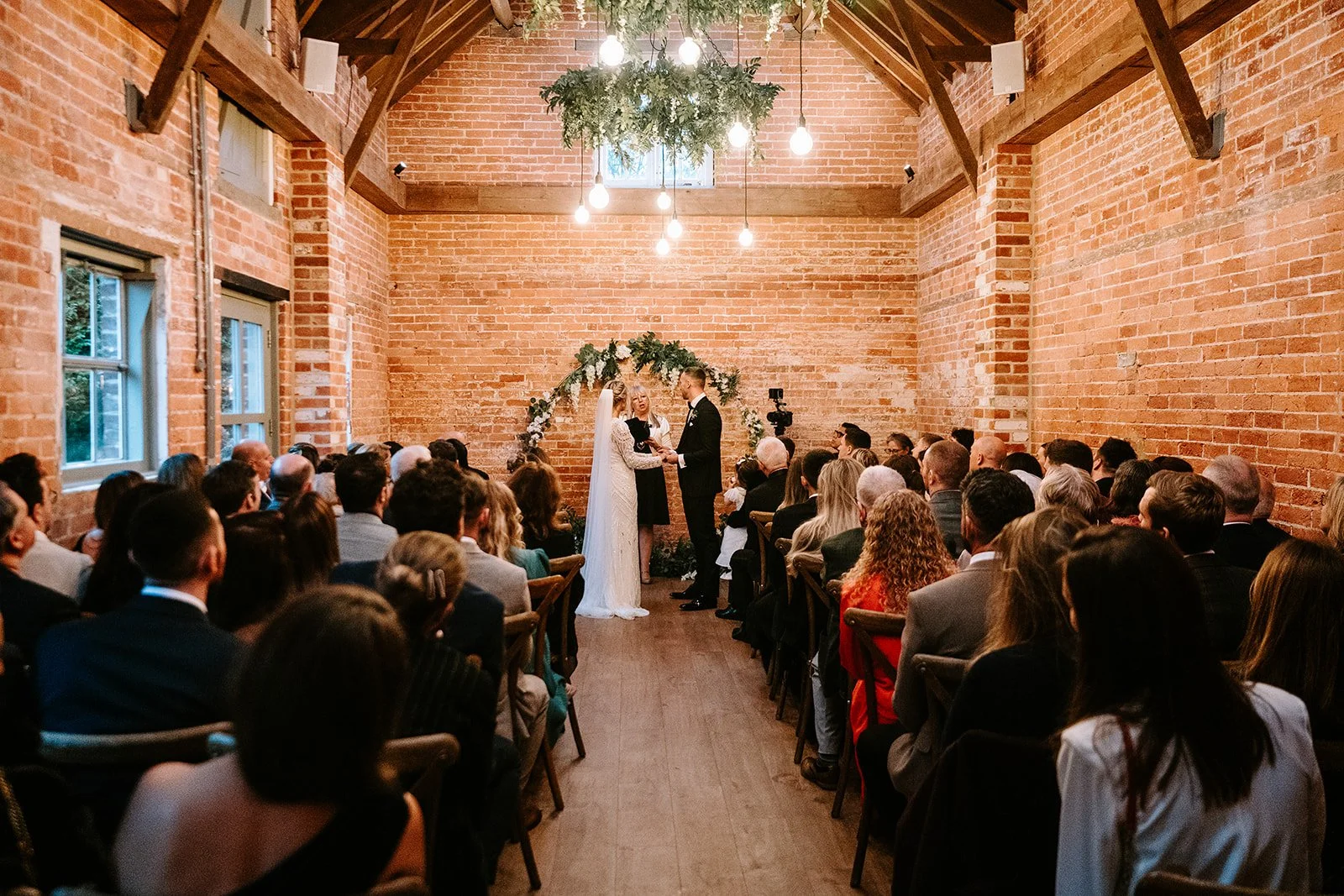 A wedding ceremony inside a rustic brick-walled hall with high ceilings, featuring a bride and groom standing at the altar. The bride wears a long white dress and veil, and the groom in a black suit. Behind them is a floral arch, and the officiant is