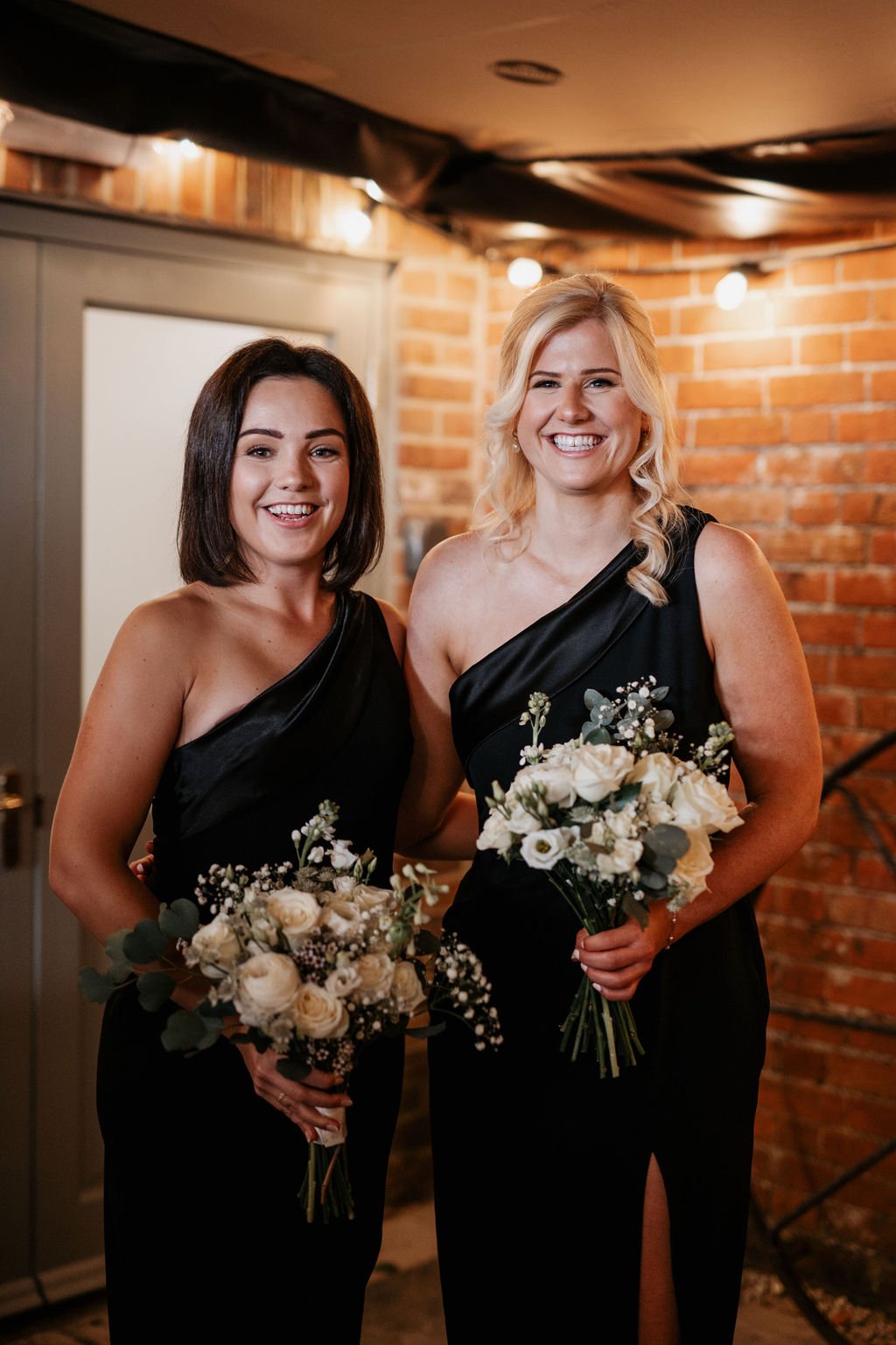 Two women in black dresses holding bouquets, standing indoors with brick wall background.