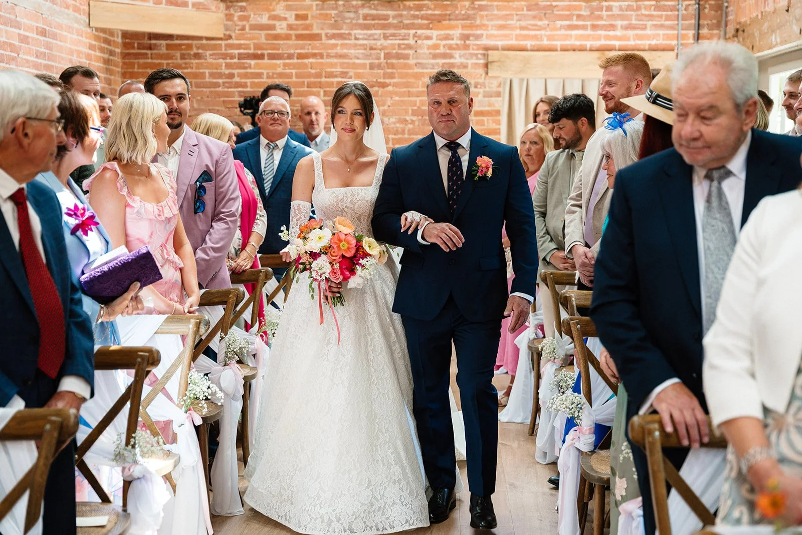A bride in a white lace wedding dress walking down the aisle arm-in-arm with her father during a wedding ceremony, surrounded by seated guests in a decorated indoor venue with brick walls.