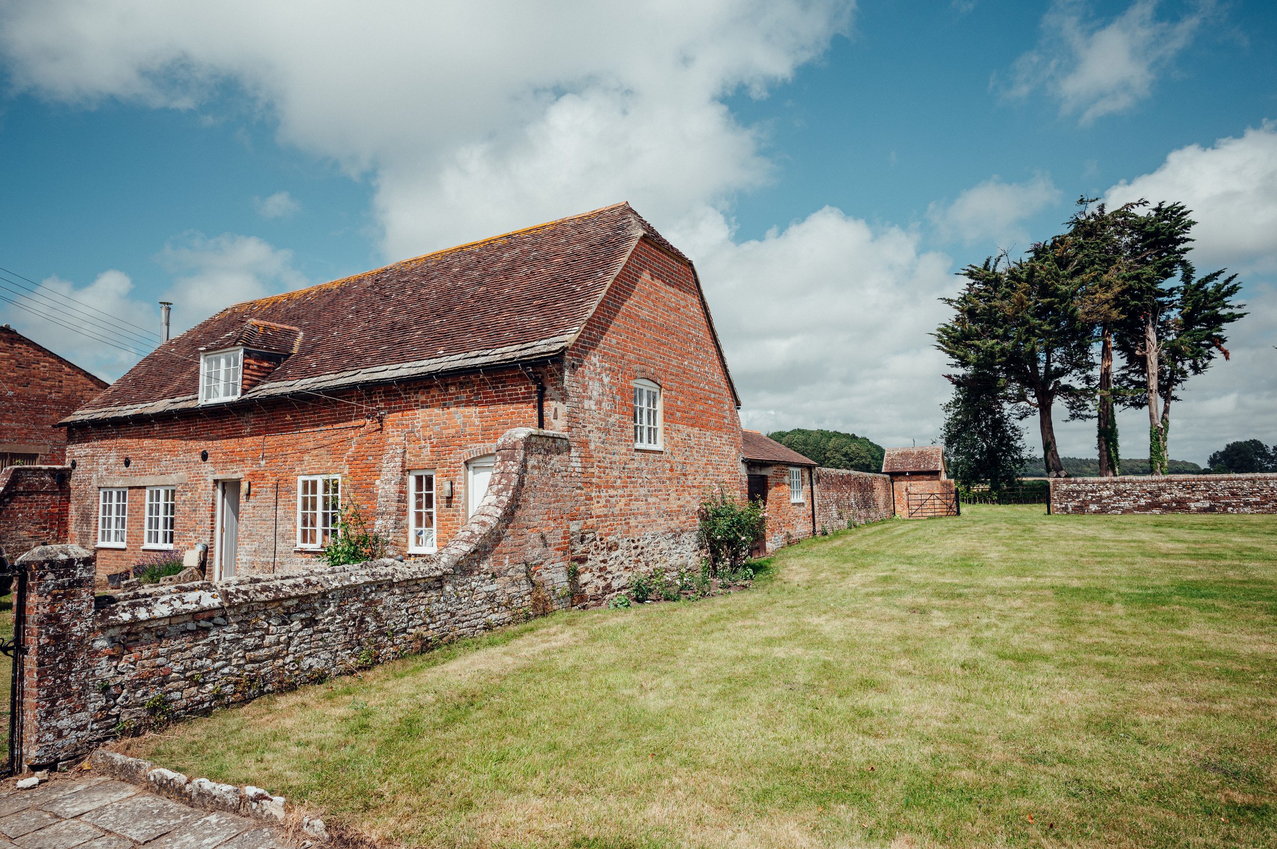 Old red brick house with slanted roof, surrounded by a stone wall, and a grassy yard with trees in the background.