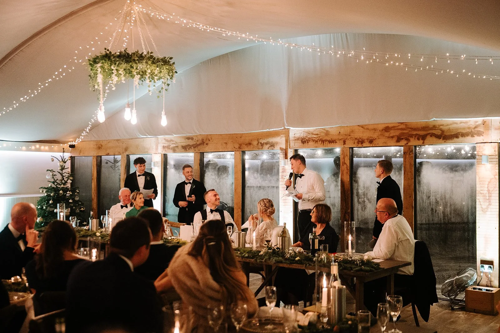 People attending a wedding reception in a decorated indoor venue with fairy lights, a Christmas tree, and a speaker giving a toast or speech.