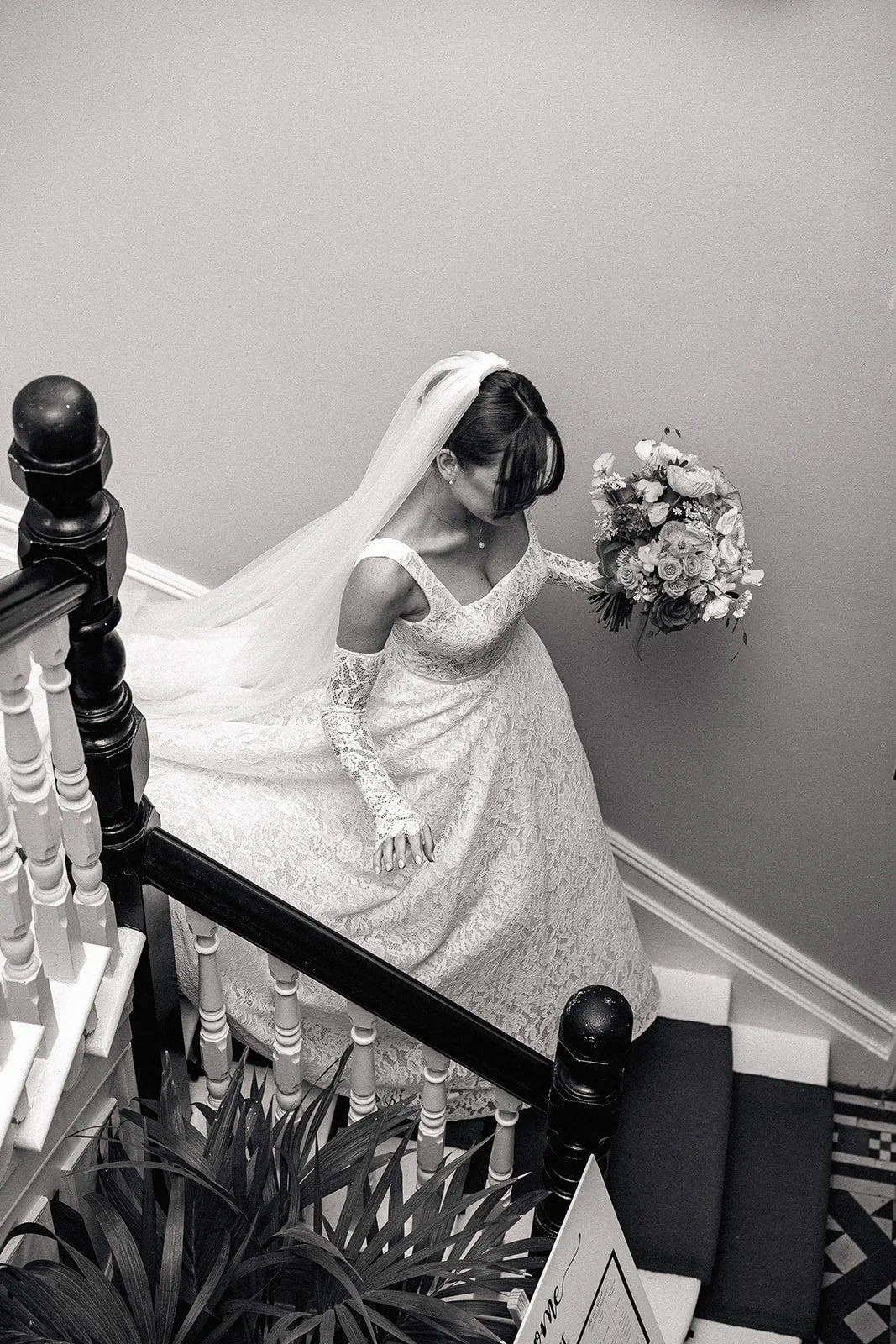 A bride in a lace wedding dress with lace sleeves, holding a bouquet of roses, standing on a staircase with a veil flowing behind her, looking down.