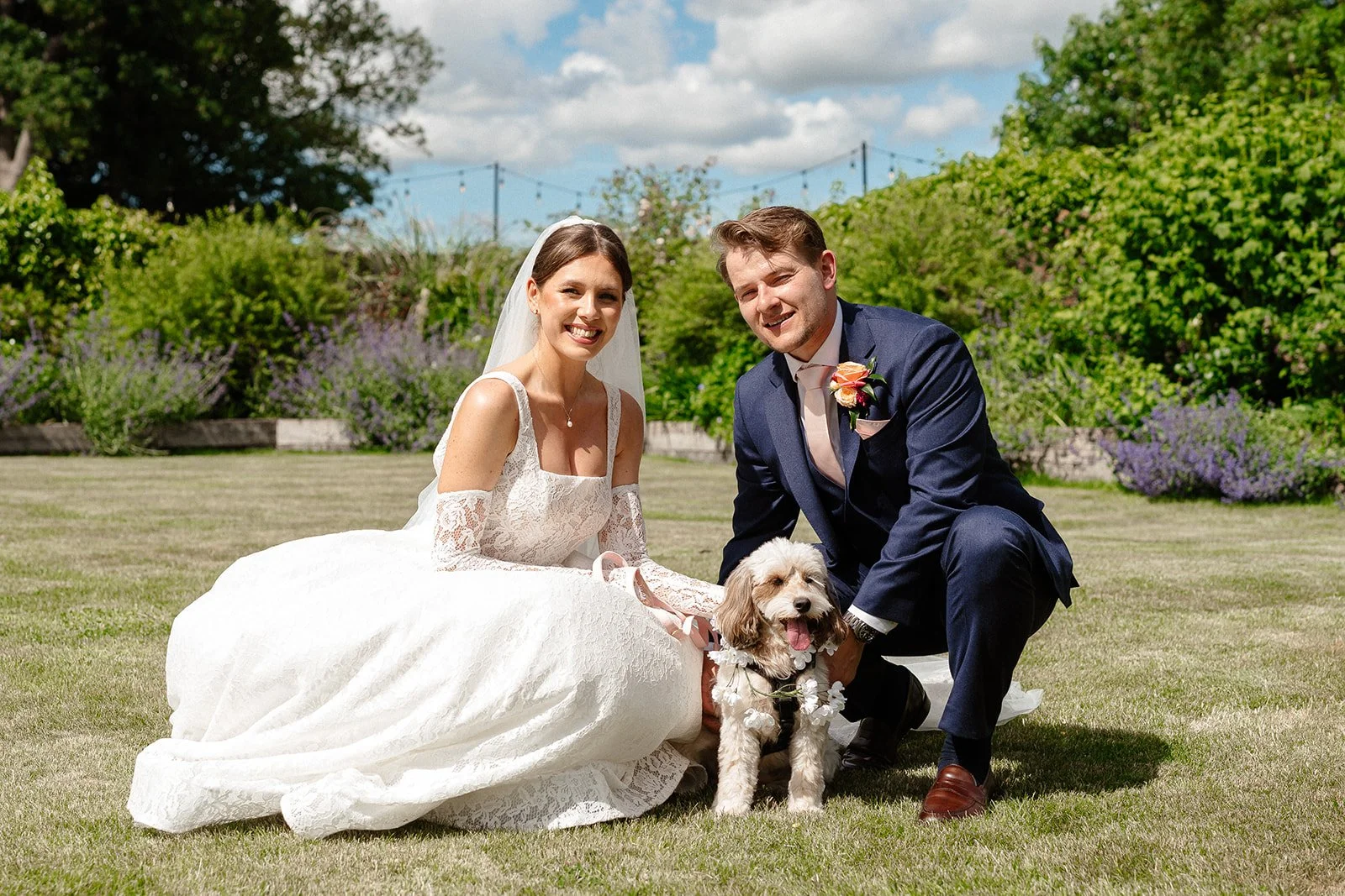 A bride and groom in wedding attire kneeling on grass outdoors with a dog between them, surrounded by green plants and trees under a partly cloudy sky.