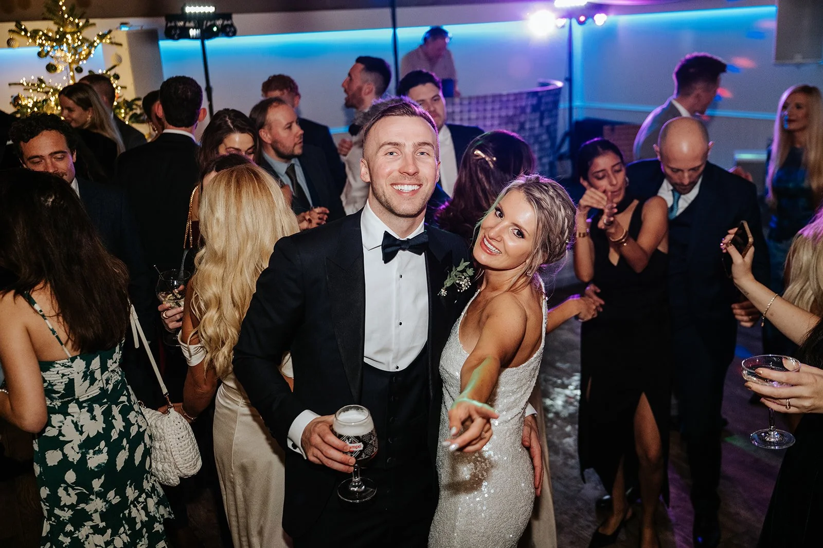 A wedding reception with a bride and groom dancing and smiling at the camera, surrounded by guests in formal attire.