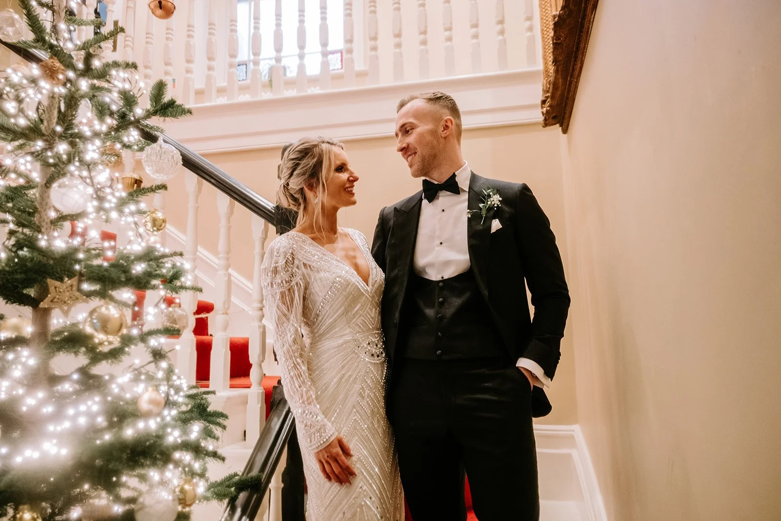 A bride and groom stand together on a staircase decorated for Christmas, smiling and looking at each other. The bride wears a white gown with intricate beading, and the groom wears a black tuxedo with a bow tie. A decorated Christmas tree with lights