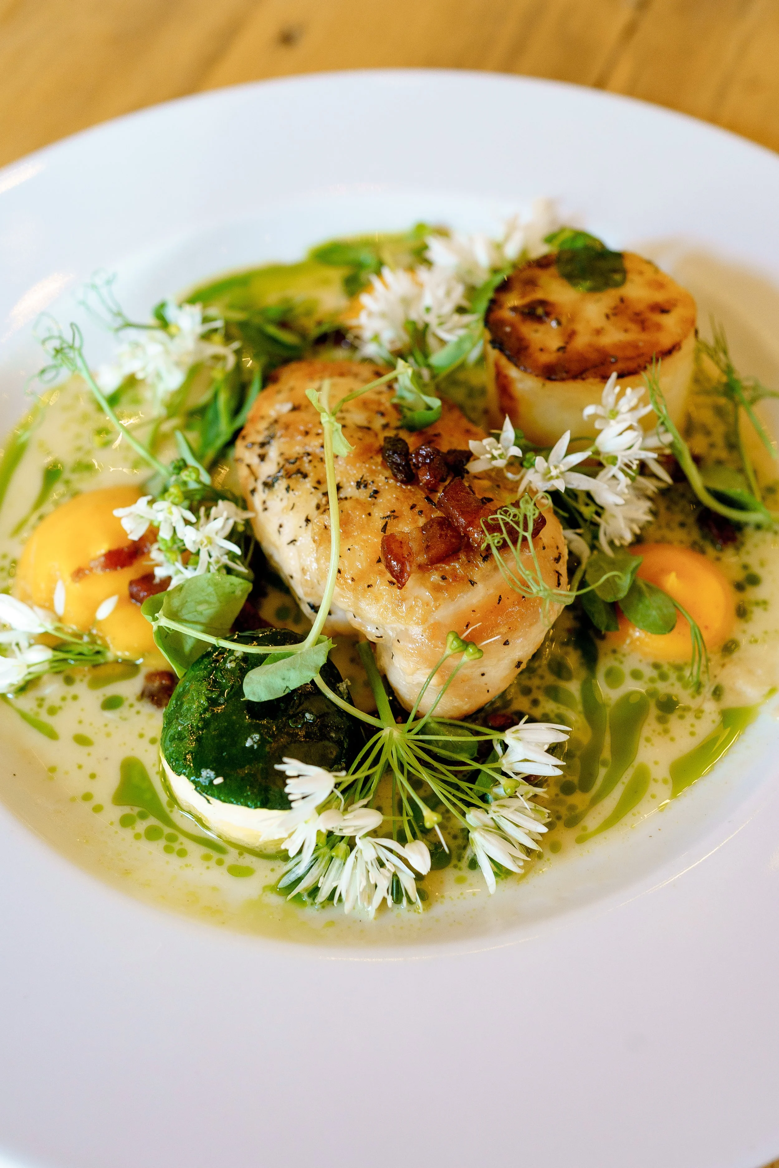 A gourmet dish featuring seared scallops, garnished with edible flowers, herbs, and green sauce, served with a lemon wedge in a white bowl.