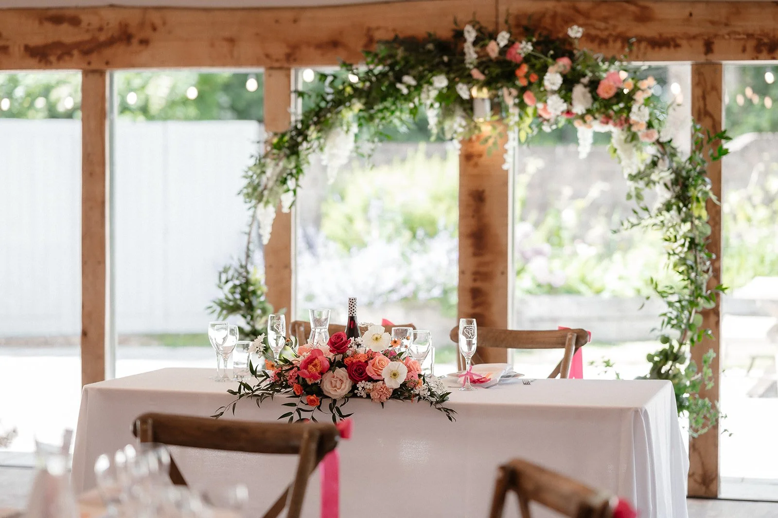 A decorated wedding table with a floral centerpiece, surrounded by wooden chairs, in a rustic venue with large windows and a floral arch