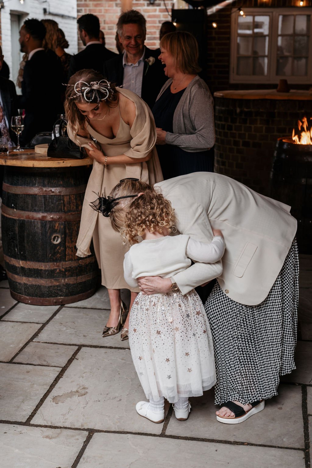 People at an indoor event with one person hugging a child in a sparkly dress, others laughing and interacting, wooden barrels in the background.