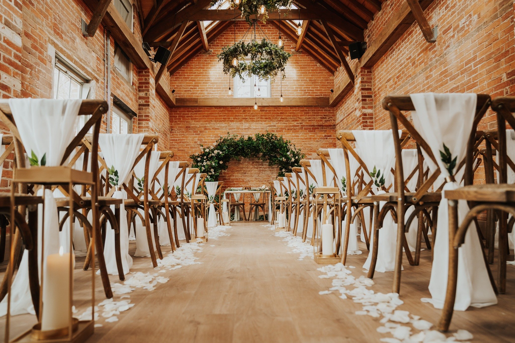 Rustic wedding ceremony setup in a brick venue with wooden chairs, white drapes, floral arrangements, and hanging greenery.