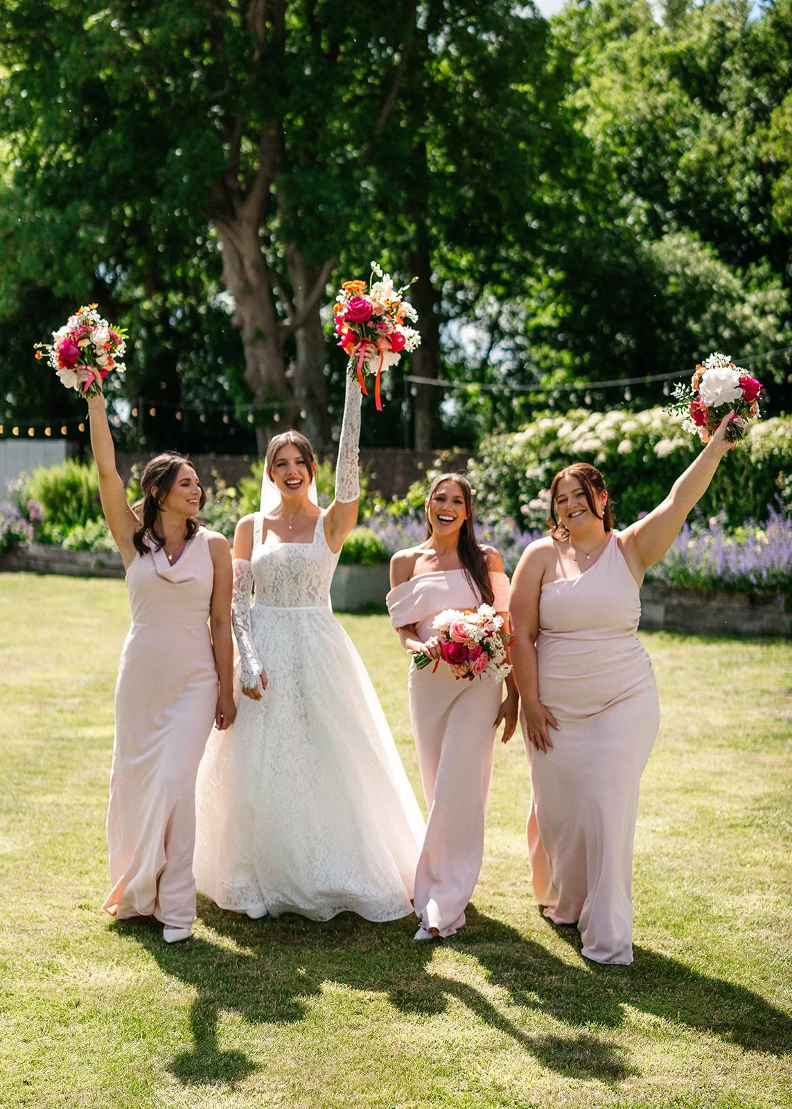 A bride and three bridesmaids celebrating outdoors on a sunny day, holding bouquets, with large trees and greenery in the background.