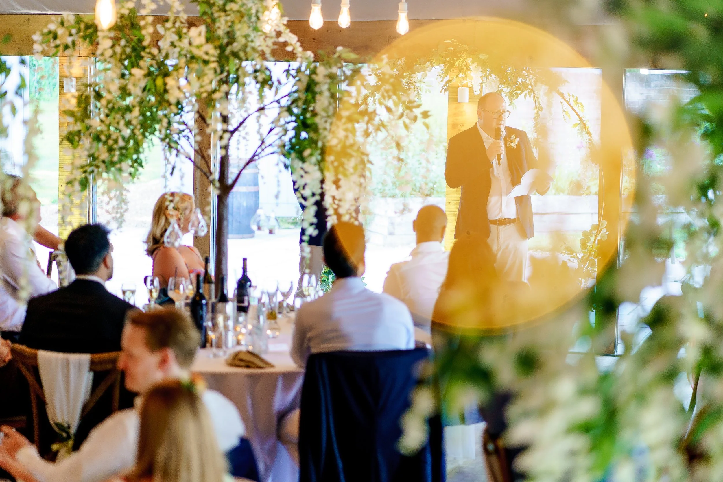 Man giving speech at wedding reception inside decorated venue with floral arrangements and seated guests.