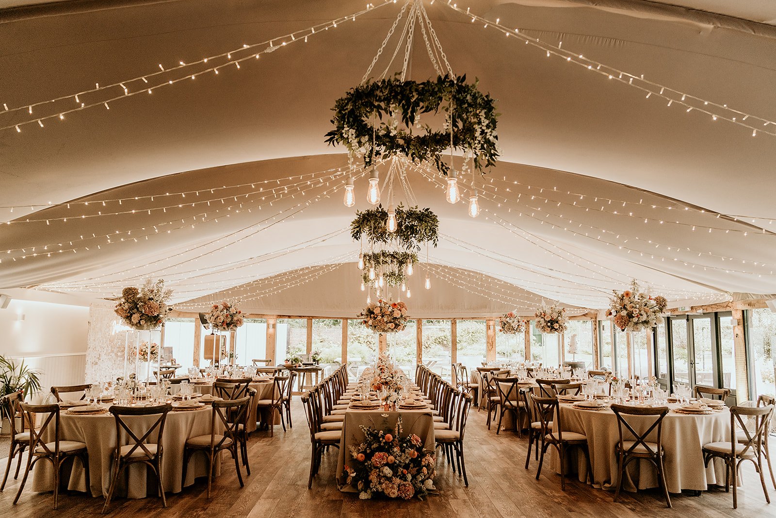 Elegant wedding reception setup with hanging lights, floral arrangements, and draped fabric in a tented venue.