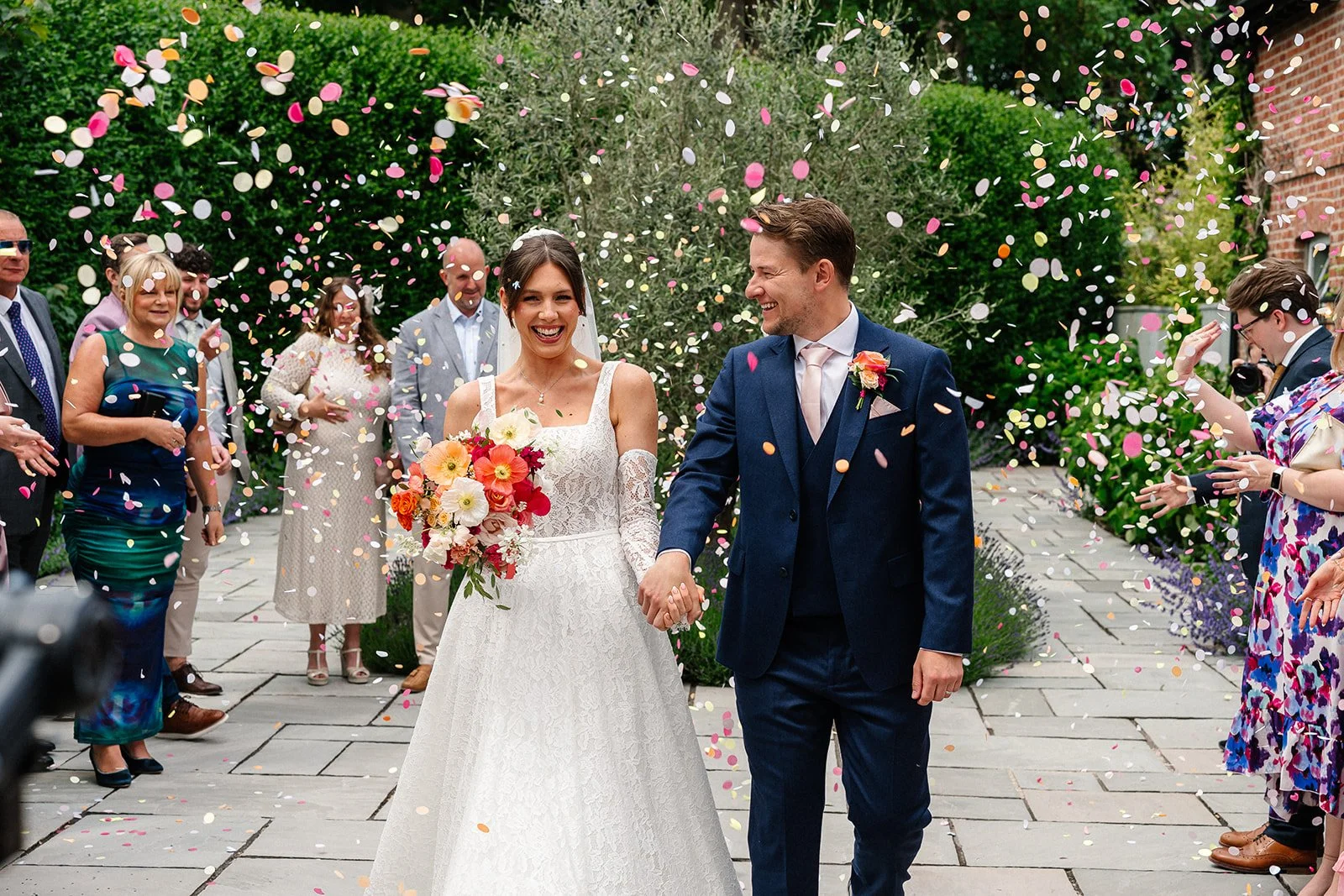 A newlywed couple is walking arm-in-arm, smiling, as colorful confetti is falling around them during their wedding celebration outdoors, surrounded by friends and family.