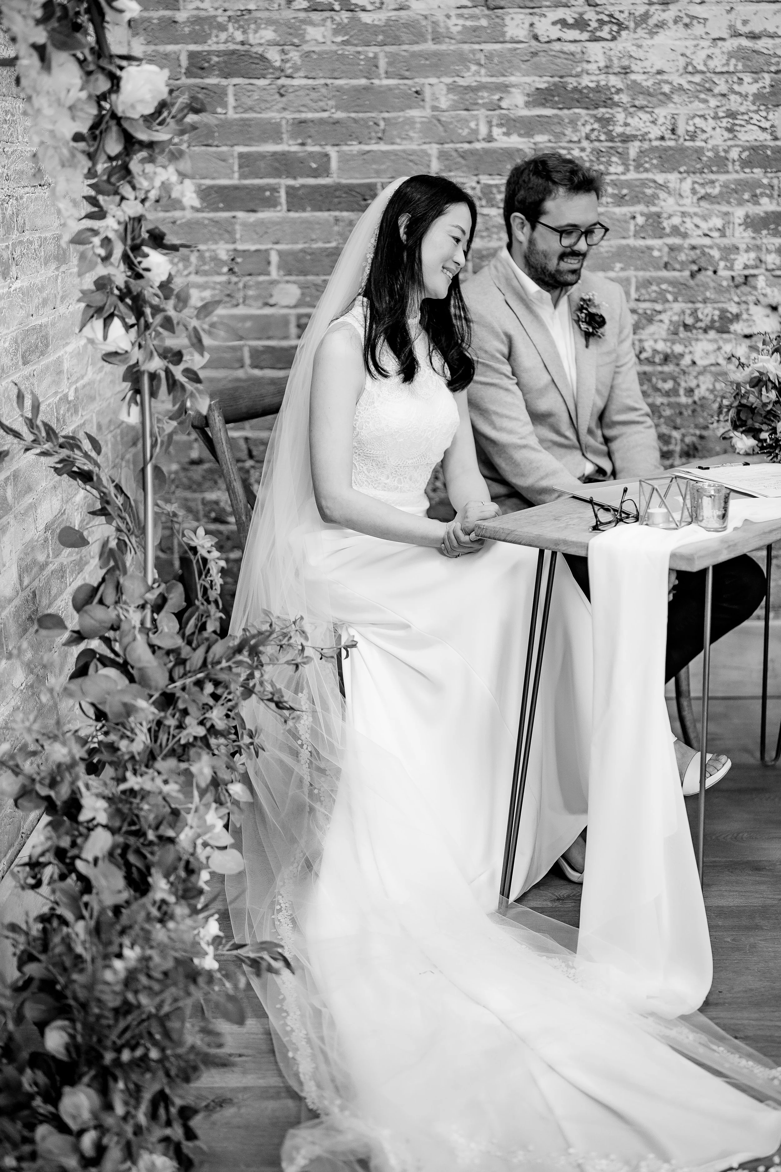 A couple at their wedding ceremony, sitting side by side, with the bride wearing a white gown and veil, and the groom in a light-colored suit with glasses, both smiling, against a brick wall backdrop decorated with flowers.