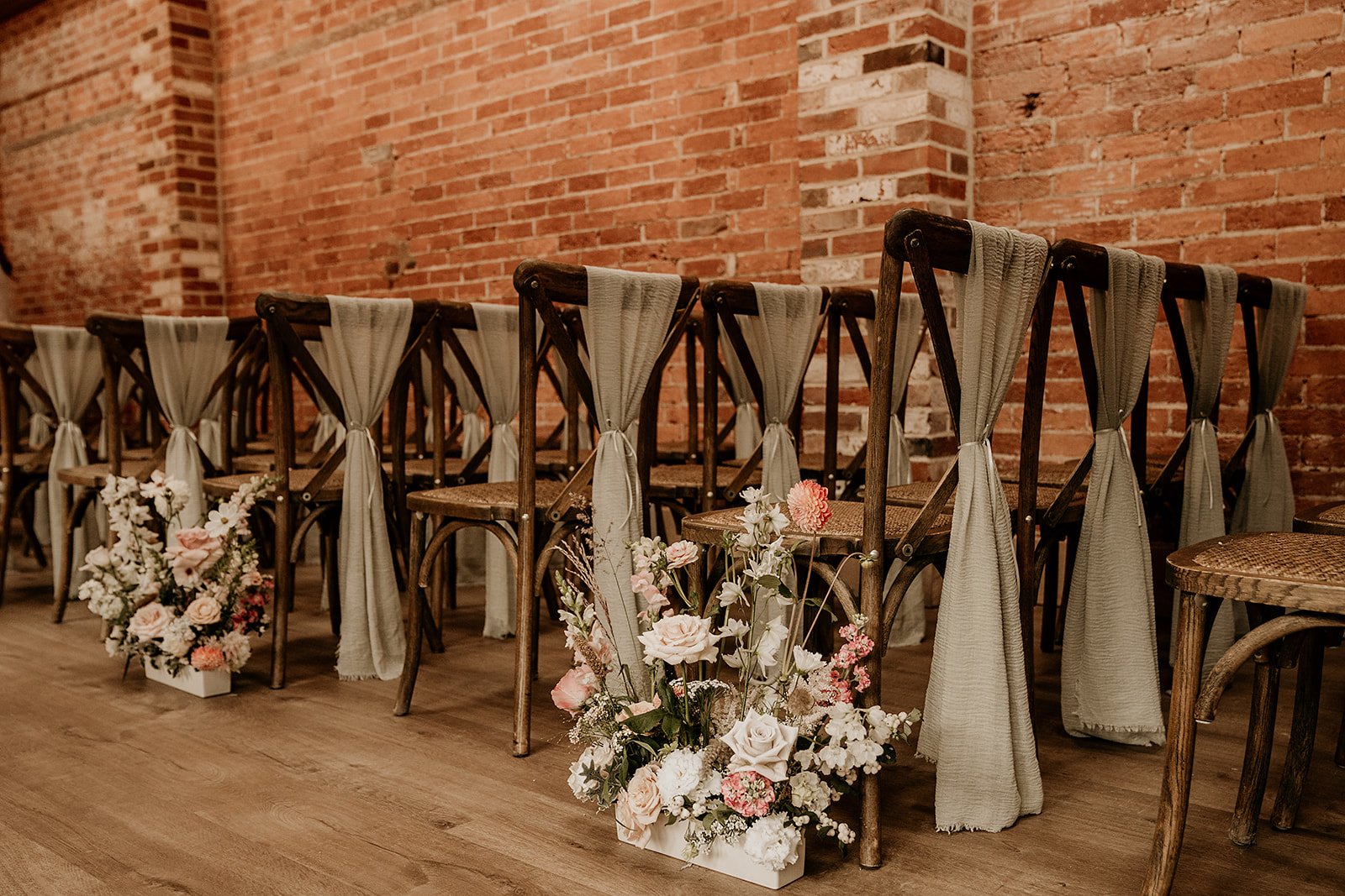 A row of wooden chairs with gray fabric draped over the backs, set in a venue with exposed brick walls. Floral arrangements with roses and greenery are placed on the floor beside the chairs.