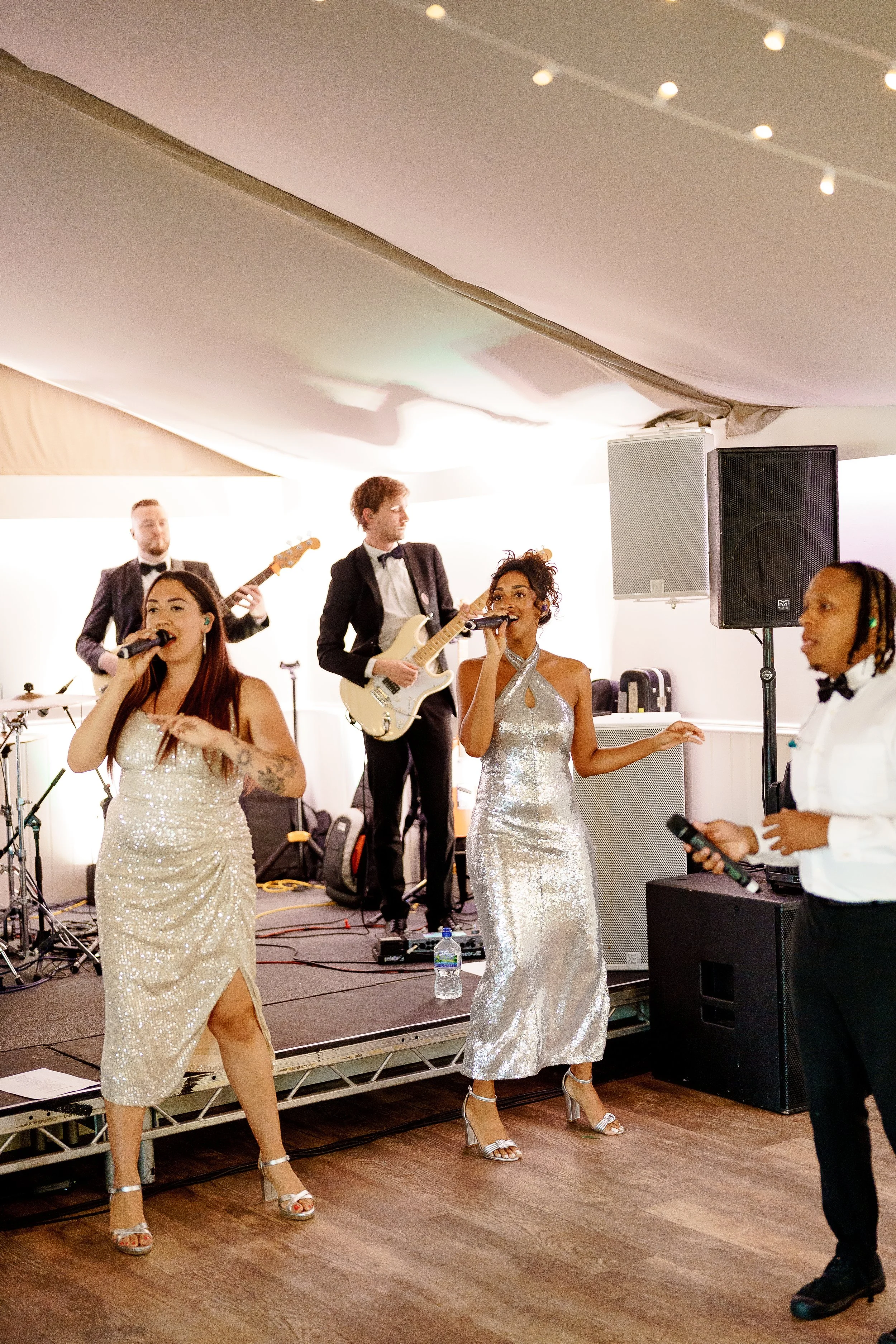 A band performing on a small stage at a celebration, with two women singing into microphones and three men playing guitar and drums in the background.