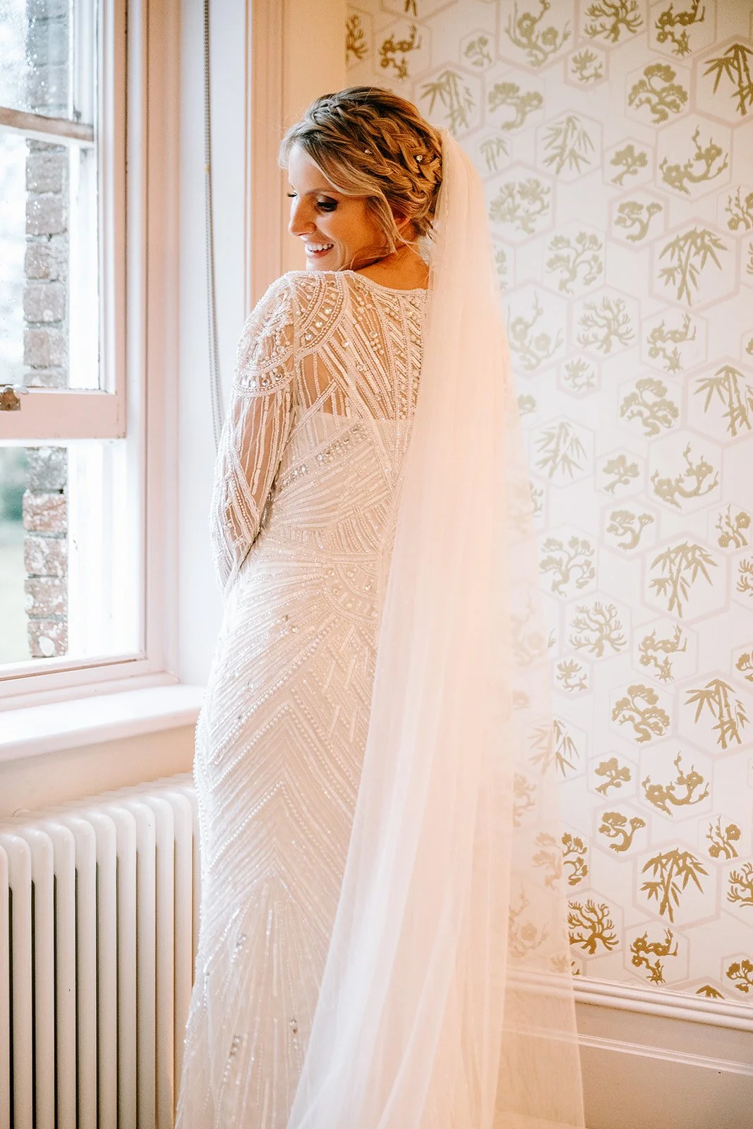 A bride in a white wedding dress with intricate beadwork, standing by a window with sunlight, smiling and looking down, with a veil cascading down her back, against a background of patterned wallpaper with gold and white designs.