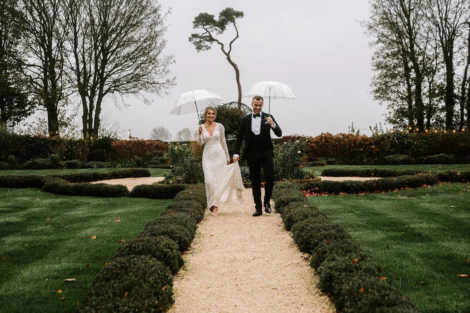 A newlywed couple walking on a garden pathway holding umbrellas on a cloudy, rainy day.