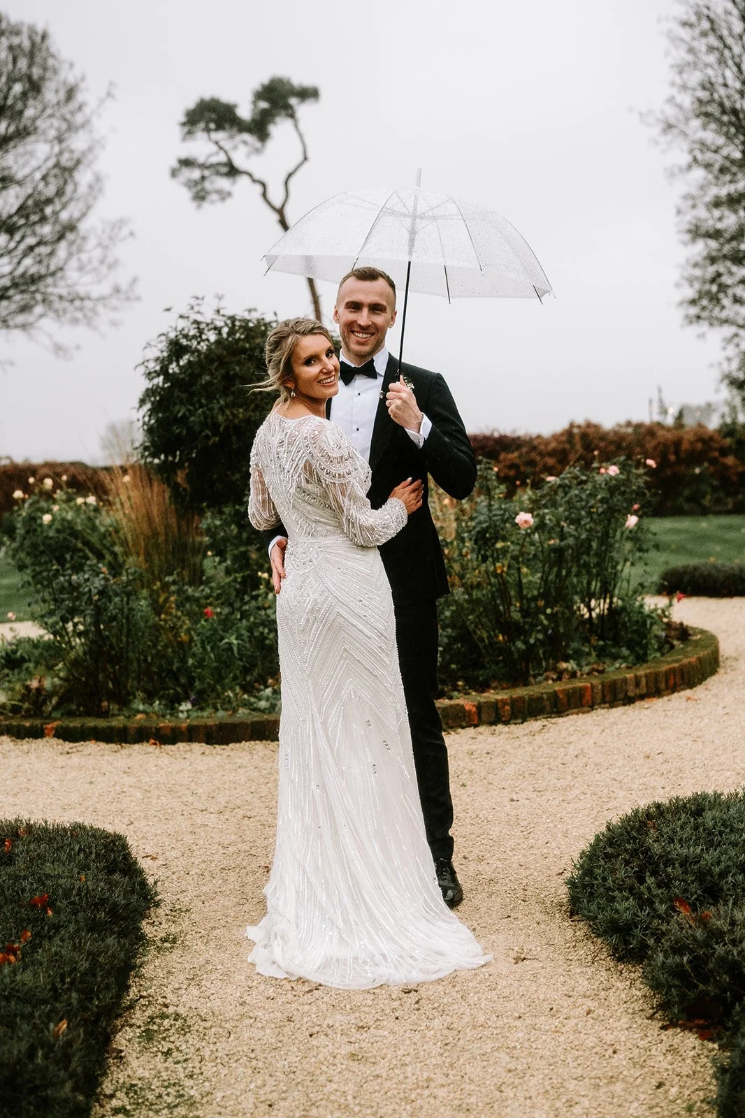 Bride and groom standing together outdoors on a gravel path with rose bushes and greenery, holding a transparent umbrella in the rain, both smiling.
