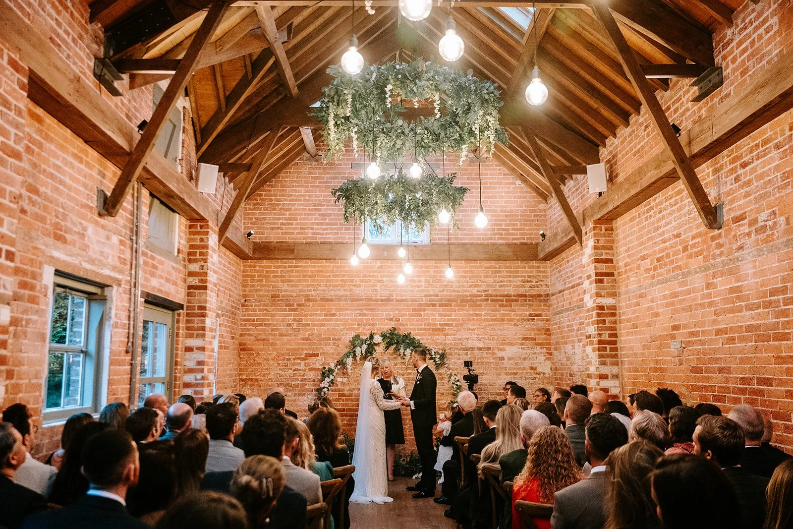 Indoor wedding ceremony in a rustic brick hall with a high wooden ceiling. The bride and groom are exchanging vows in front of a floral arch, with guests seated on wooden benches watching.