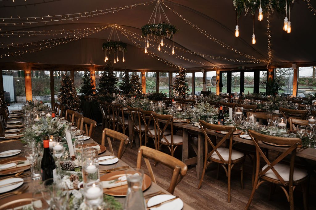 Elegant banquet hall decorated for an event with wooden tables, candles, greenery, and festive lights. Large windows and Christmas trees are visible in the background.