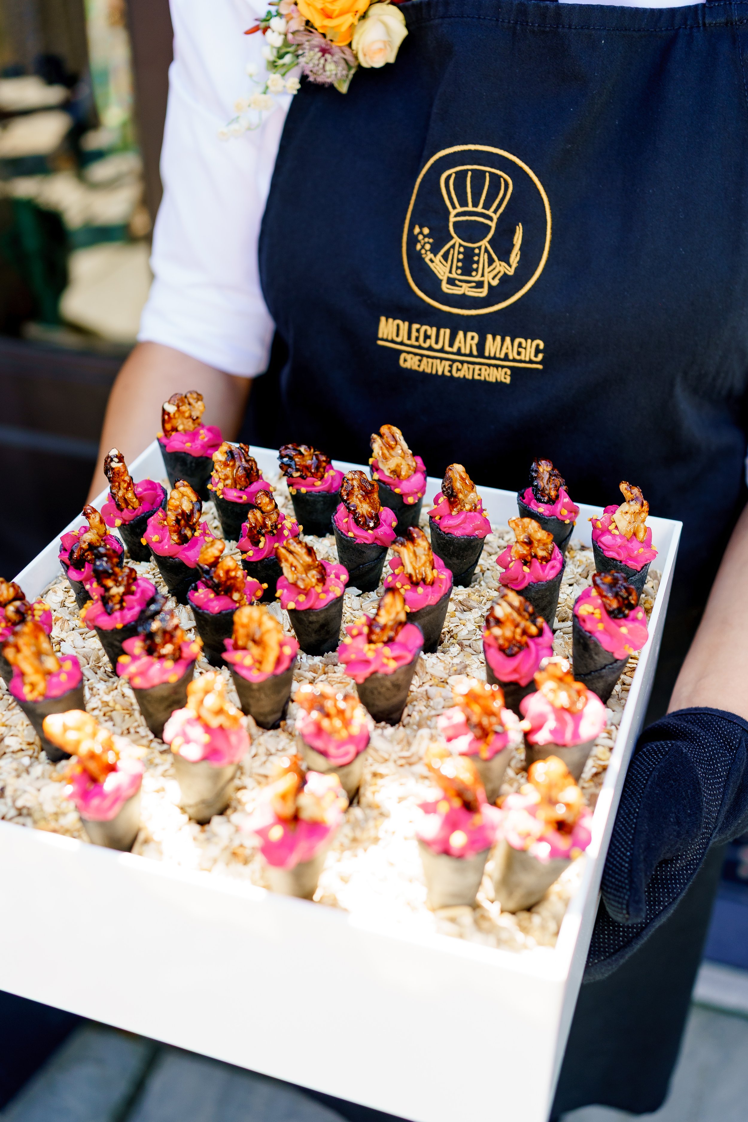 A person wearing a black apron with 'Molecular Magic Creative Catering' written on it is holding a white tray filled with small, cone-shaped appetizers topped with pink sauce and garnished with walnuts and yellow sprinkles. The person is also wearing