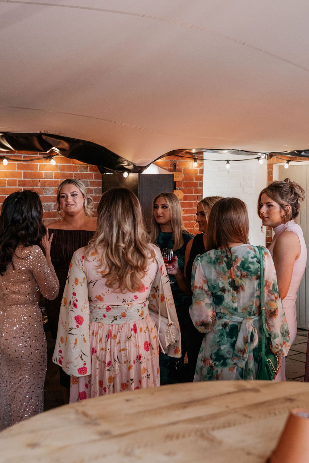 A group of women in dresses talking in a warmly lit room with a brick wall and string lights.
