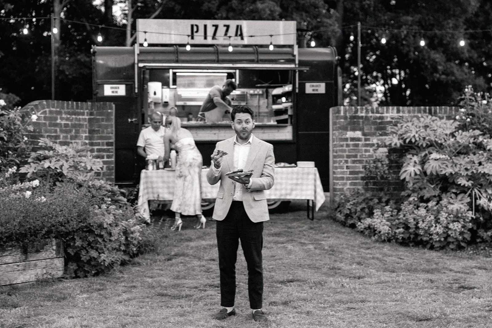 A man in a light-colored blazer holding a plate of food and a drink, standing outdoors in front of a food truck labeled 'PIZZA', with two people serving behind the counter.