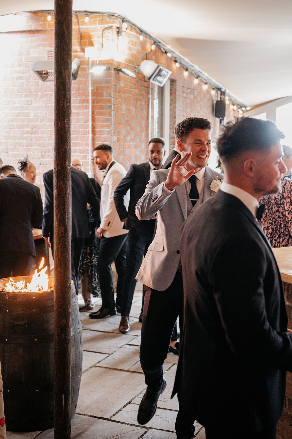 A group of people dressed formally at a social event indoors, with one person in a light suit waving. A small fire burns in a barrel near them, against a backdrop of exposed brick walls with string lights.
