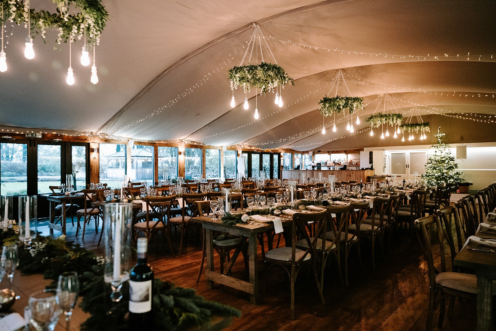 Wedding reception hall decorated with hanging lights, greenery, and a Christmas tree, with wooden tables set with glassware and silverware.