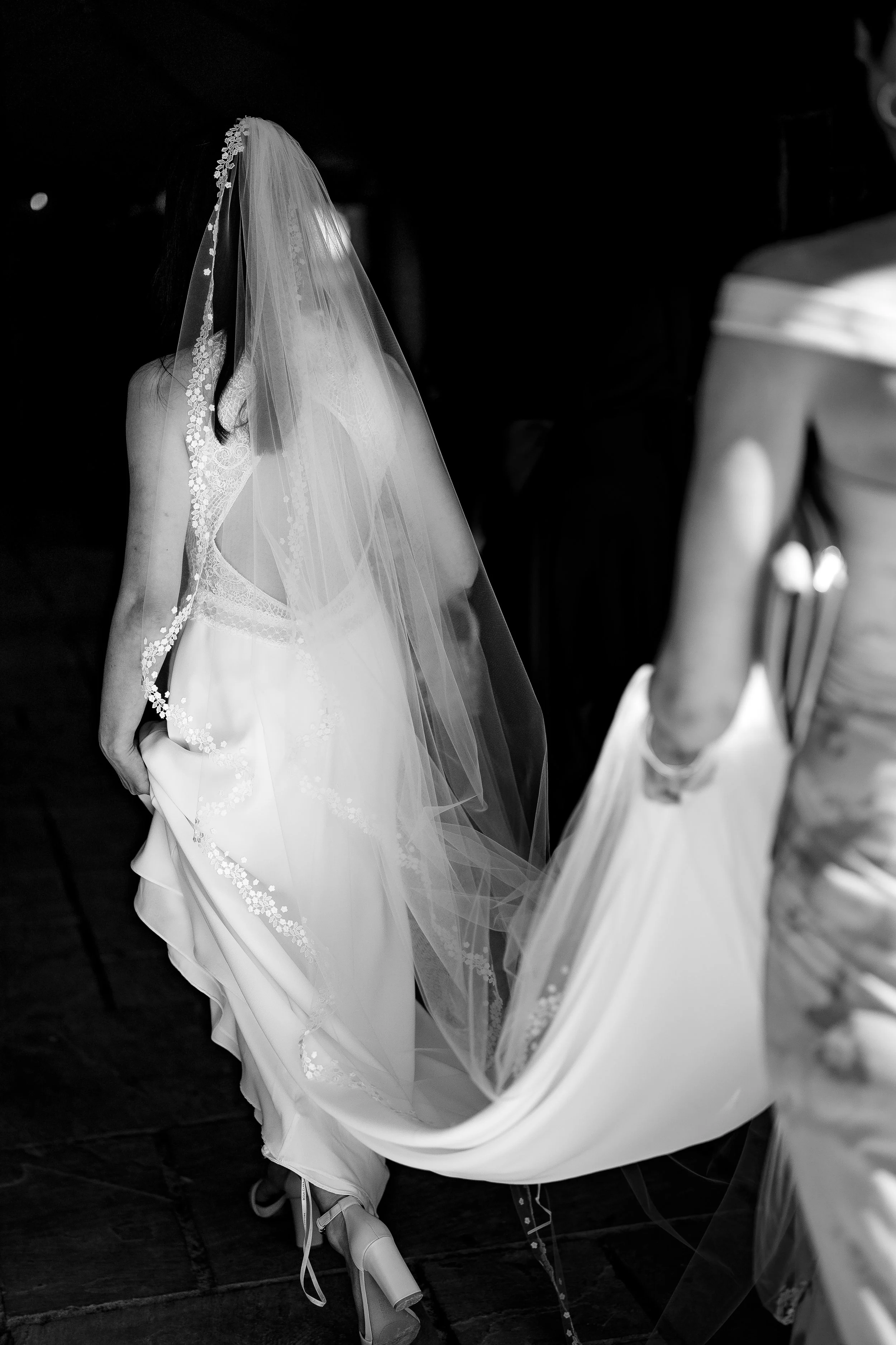 A bride in a wedding dress with a veil, holding the train of her dress as she walks, seen from behind in a black and white photo.