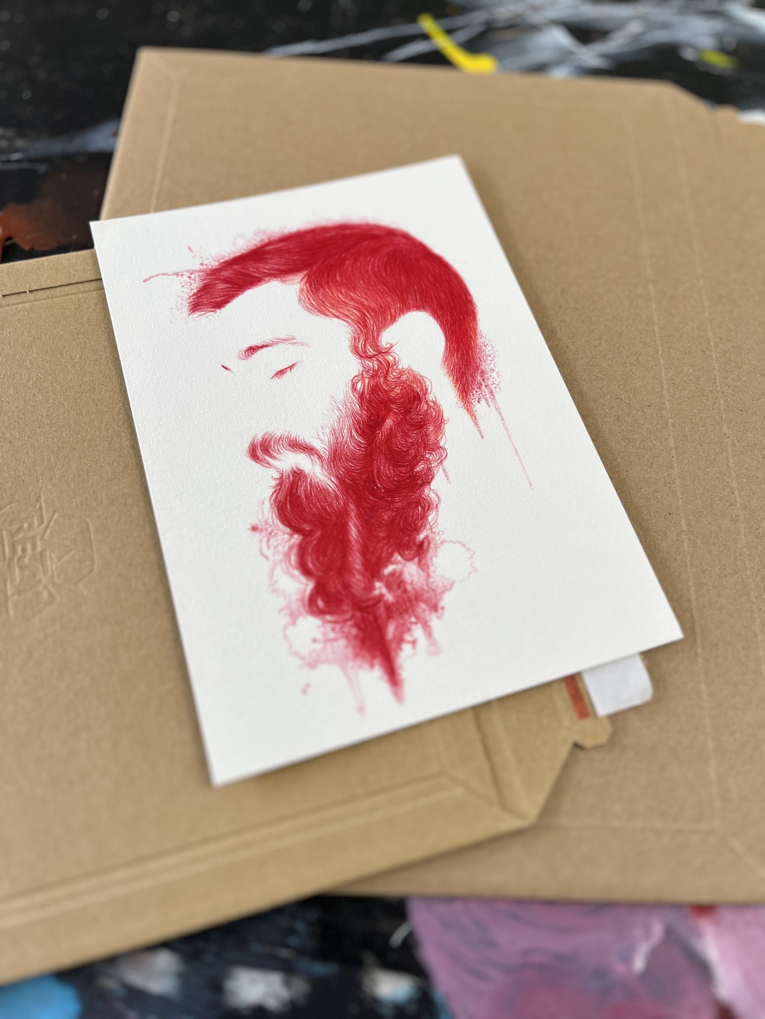 un dessin au stylo rouge représentant un homme barbu avec des cheveux longs, vue de profil. Le dessin est posé sur un fond d'enveloppe en  carton kraft.