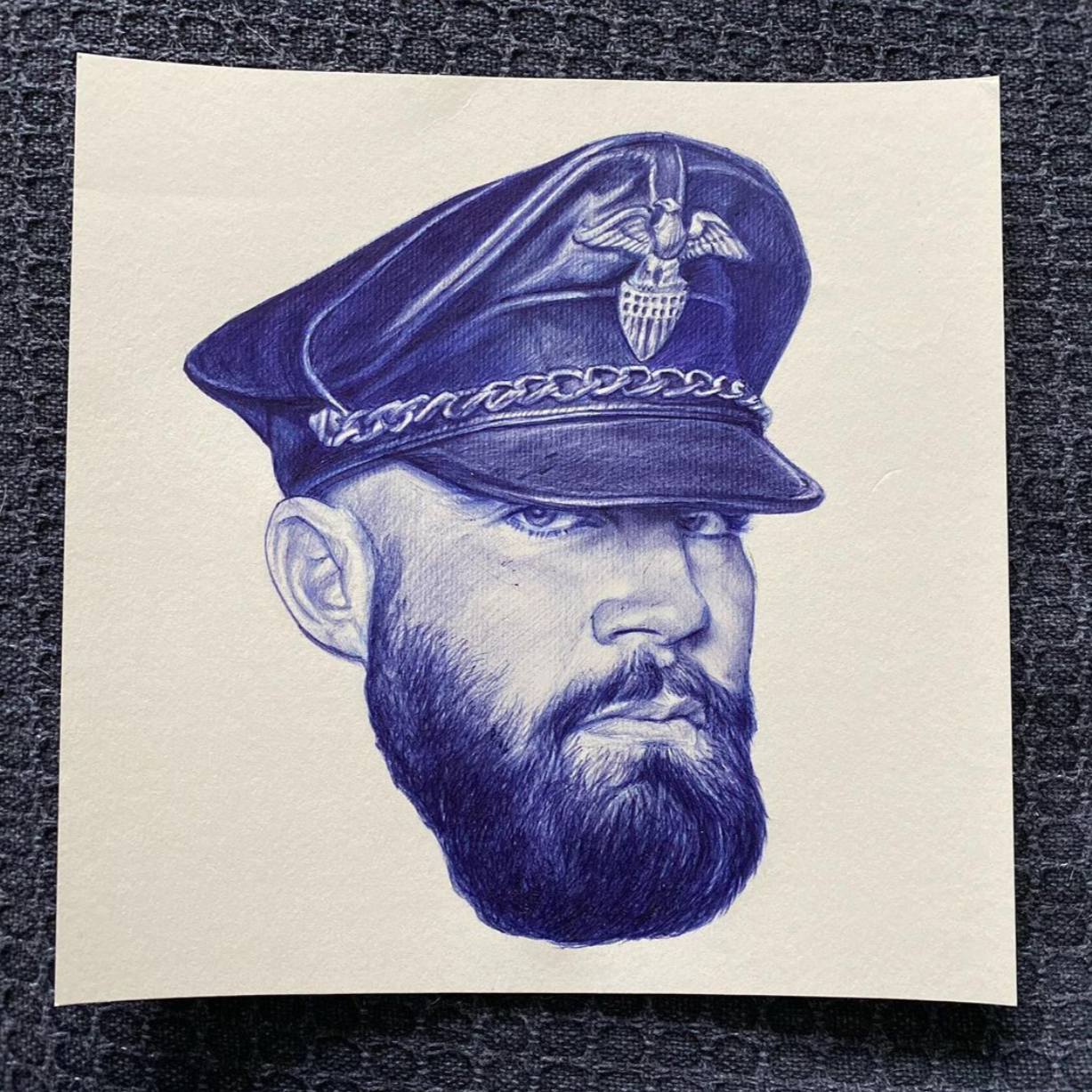 Un dessin en bleu, au stylo,  d'un homme barbu, portant une casquette en cuir avec un badge.