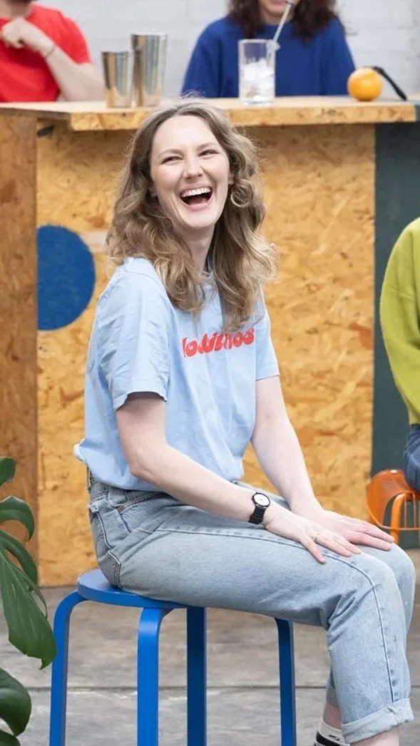 A woman with wavy light brown hair smiling and laughing, sitting on a blue stool in an indoor setting with a wooden bar in the background. She is wearing a light blue t-shirt and light jeans.