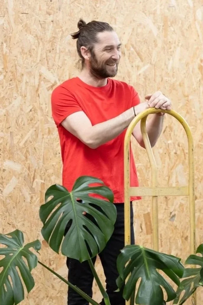 A man with long hair tied in a bun, wearing a red t-shirt and black pants, smiling and leaning on a yellow hand truck. In front of him, there are green monstera leaves, and he is standing against a wooden wall background.