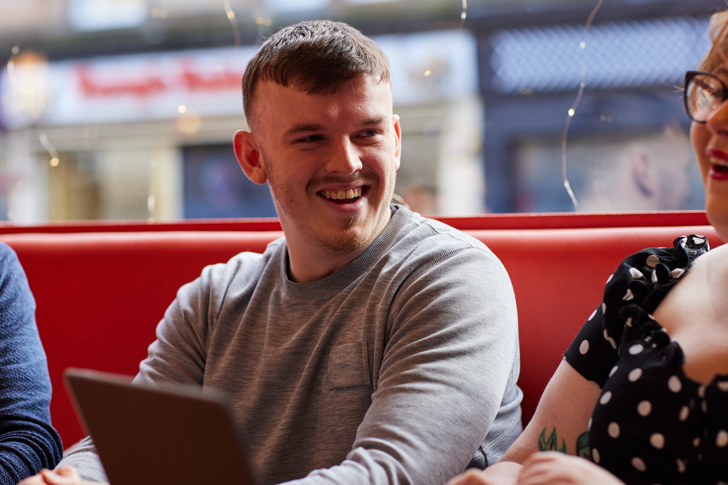 A young man with short brown hair and a goatee smiling, wearing a gray long-sleeve shirt, sitting at a table with a woman wearing glasses and a black polka dot dress, in a restaurant or cafe.