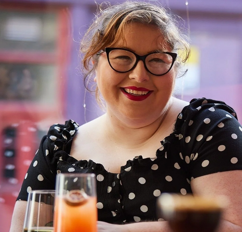 A woman with glasses and red lipstick smiling at a table with drinks, wearing a black polka dot dress with ruffled sleeves.