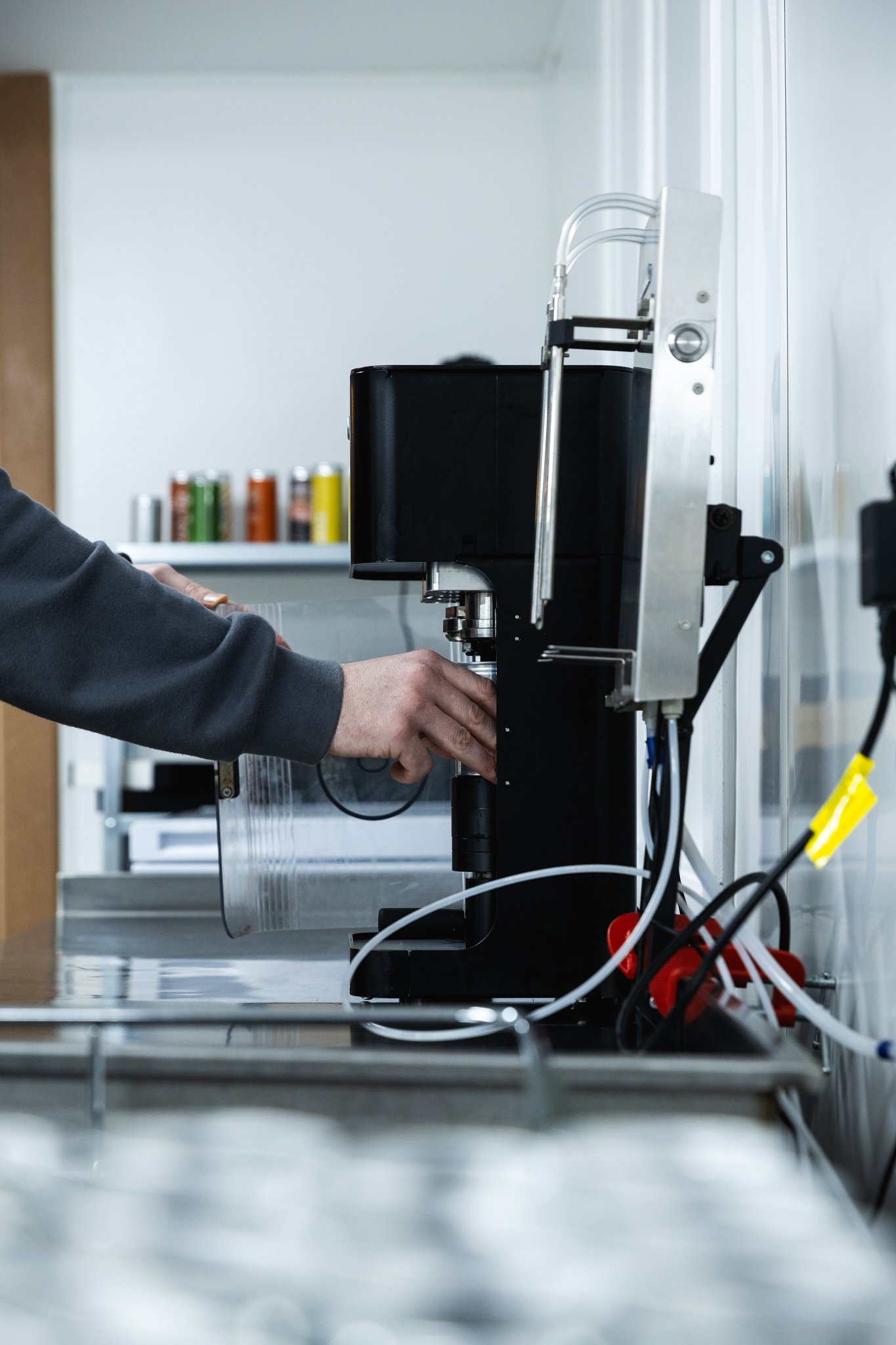 Person operating a black cocktail canning machine in a drinks lab on a stainless steel table, with a white wall and colourful cans or containers in the background.