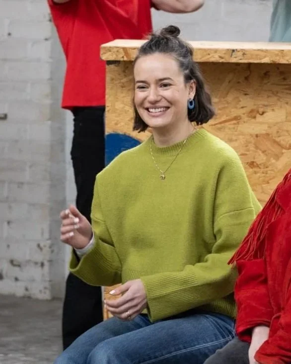 A woman with short dark hair smiling, wearing a bright green sweater, sitting in a casual setting with a wooden table behind her.