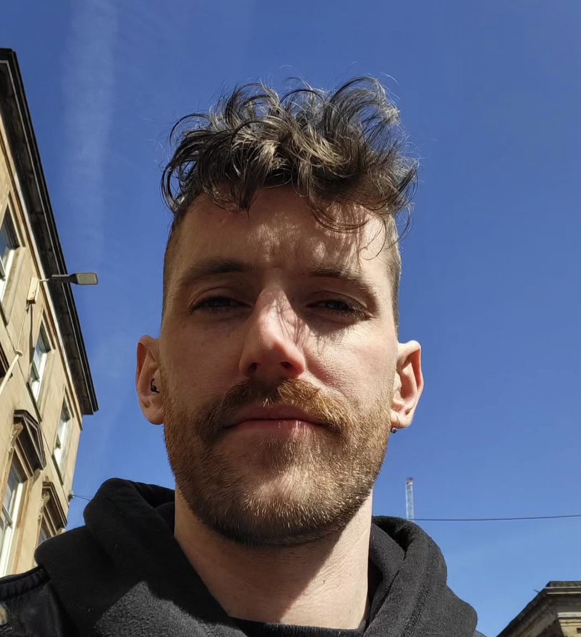 A young man with curly hair and a beard taking a selfie outdoors against a blue sky, with buildings in the background.