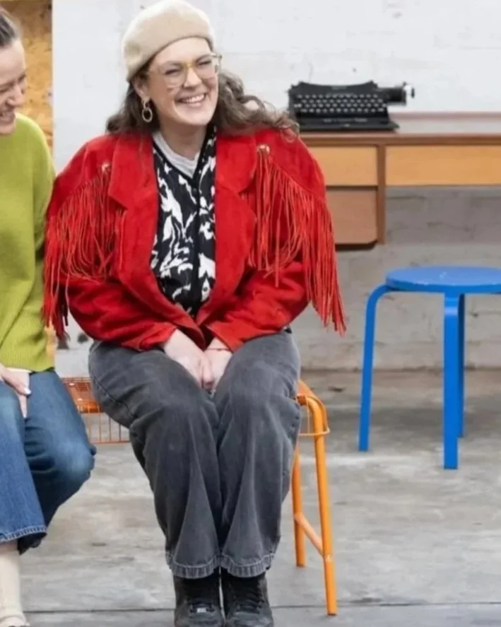 A woman with curly hair wearing glasses, a beige beret, and a red jacket with fringe details, smiling and sitting on an orange chair in a room with a typewriter on a desk and blue stool.