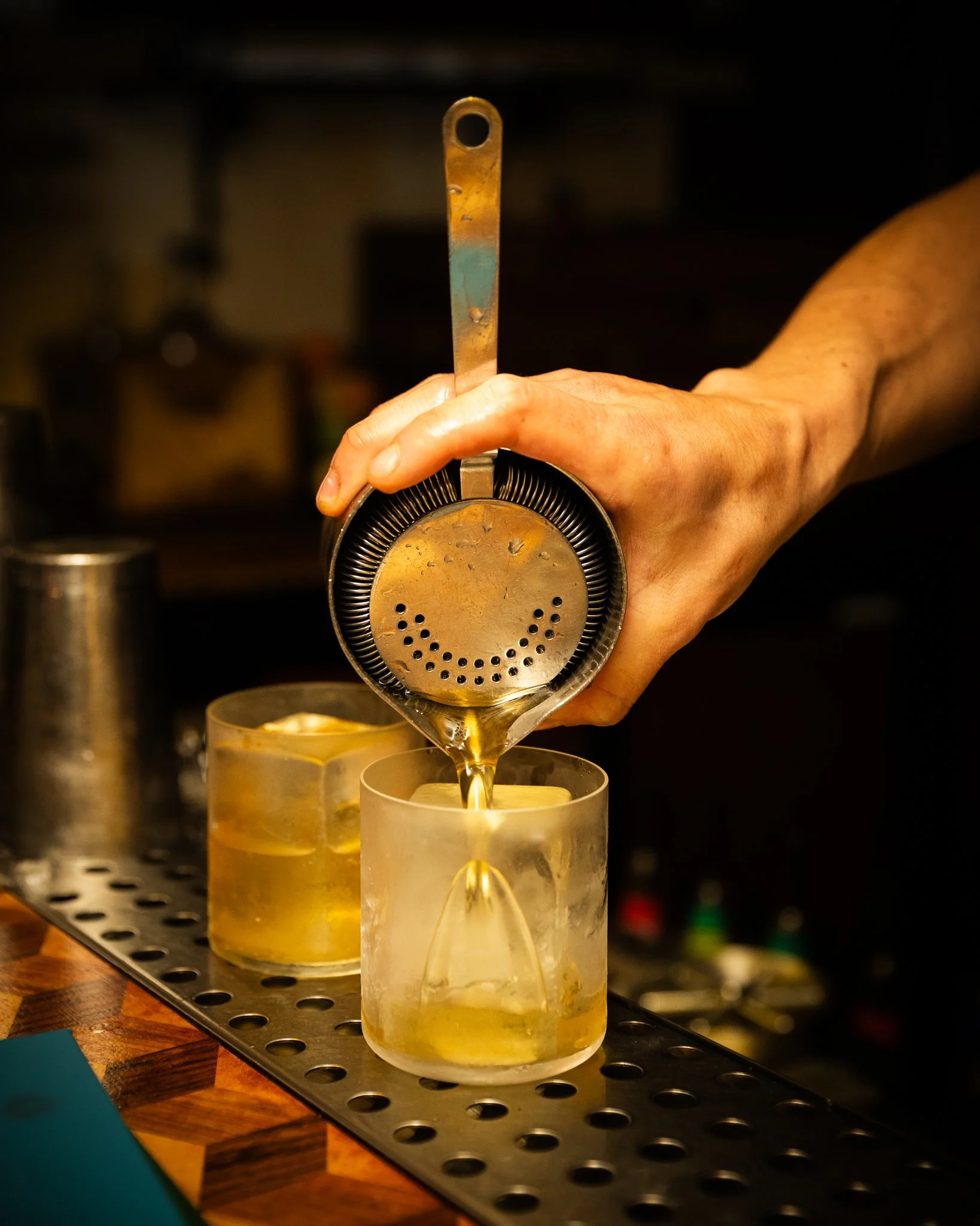 A person pouring a cocktail through a shaker strainer into a glass with ice and lemon slices behind on a bar counter.