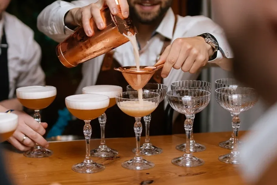 Bartender pours a pink cocktail through a sieve into coupe glasses, with several other cocktails on the wooden bar counter.
