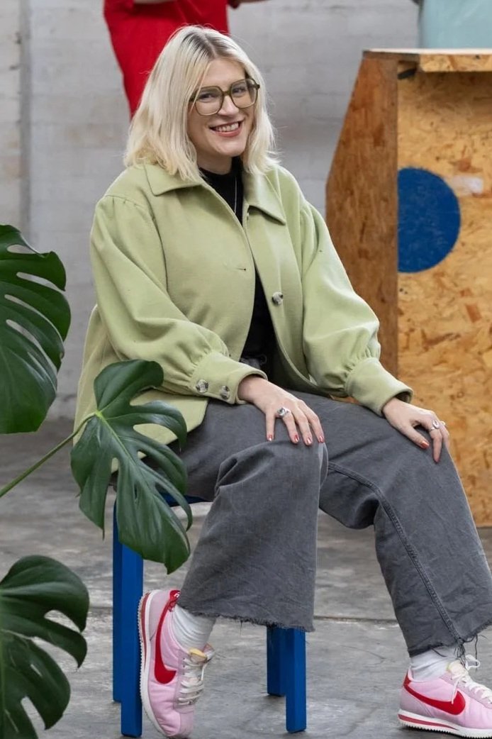 A woman with blonde hair and glasses smiling while sitting on a blue stool. She is wearing a light green jacket, dark gray wide-leg jeans, pink and white sneakers, and white socks. A large green plant partially covers her in the foreground, and a wooden object with a blue circle is in the background.