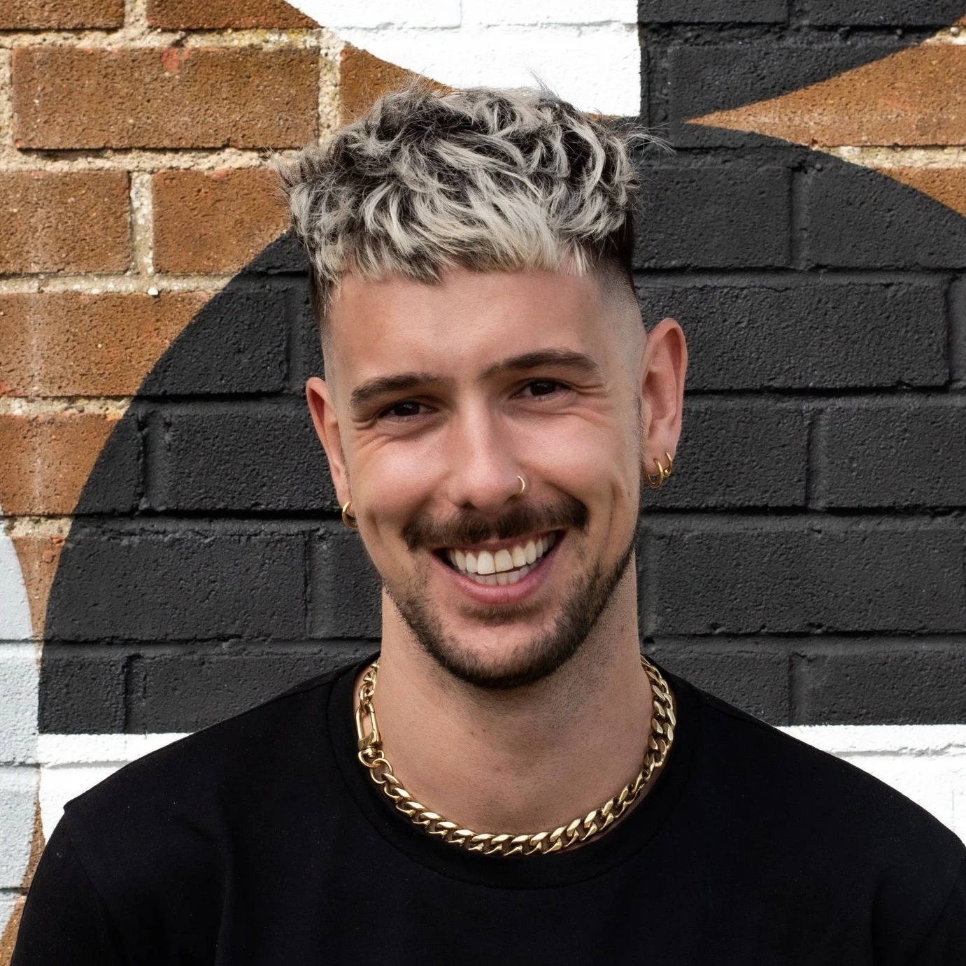 A smiling man with blond, curly hair and tattoos, wearing gold jewelry, standing in front of a brick wall painted with black and white abstract designs.