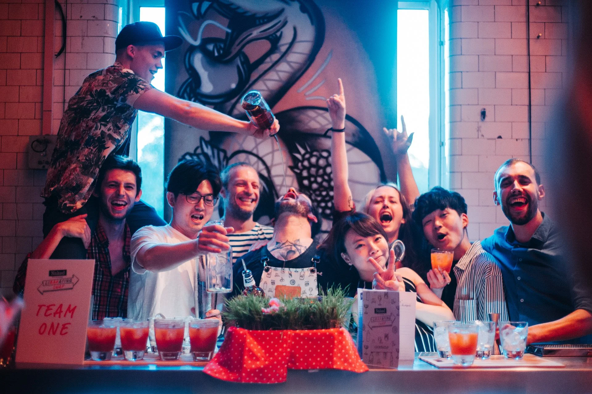 A group of people celebrating at a party around a table with drinks, smiling and having fun, with a dragon mural on the wall behind them.