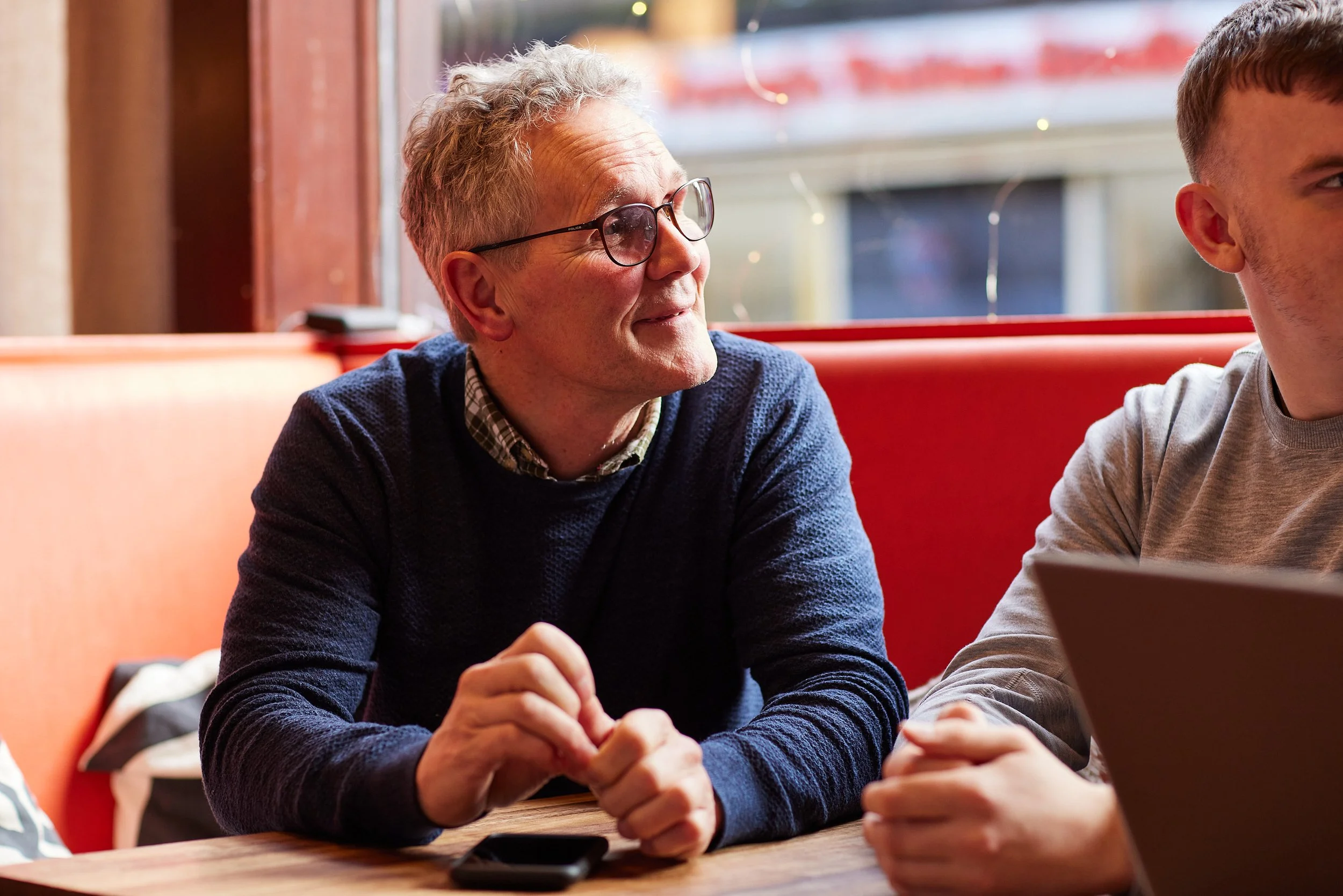 Older man with glasses and gray hair smiling and engaging in conversation with a younger man at a cafe table.