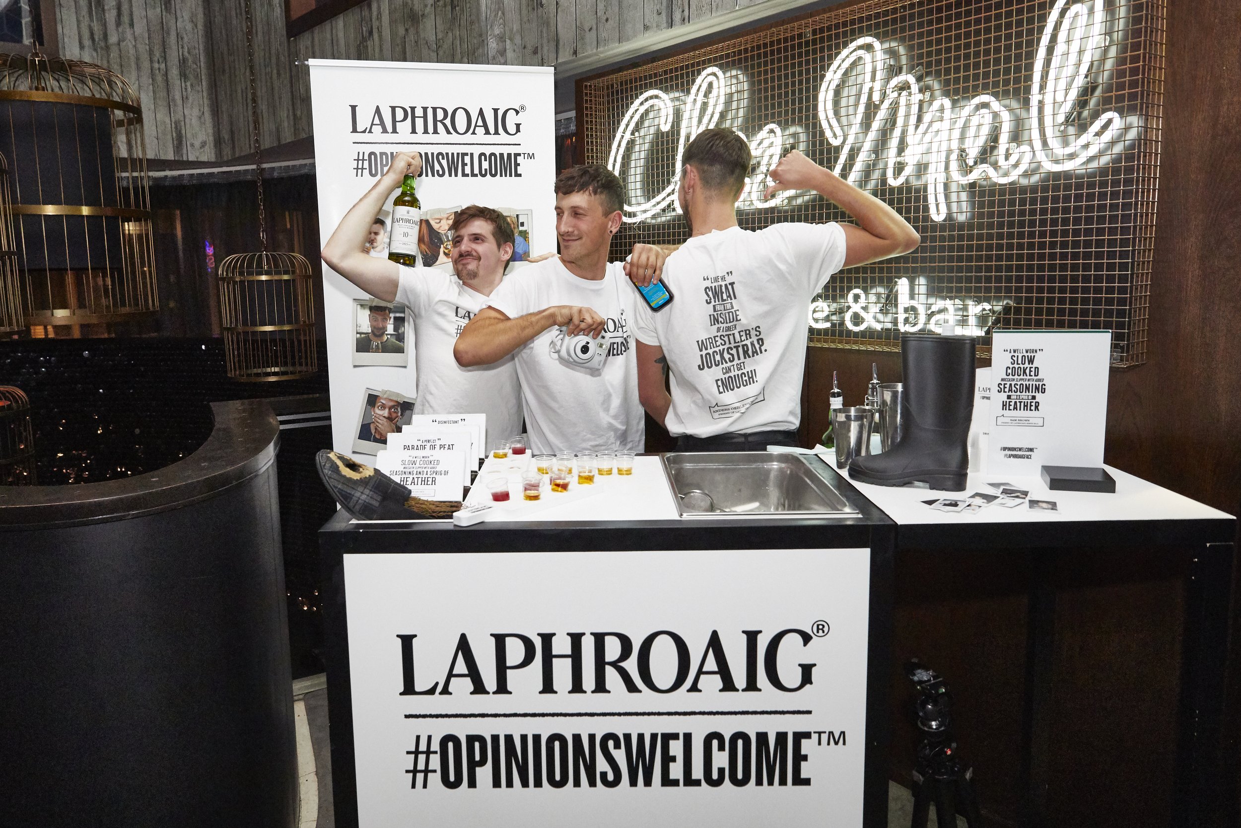 Three men standing behind a bar counter with promotional signs and decorations, celebrating with drinks and taking photographs at an event for Laphroaig.